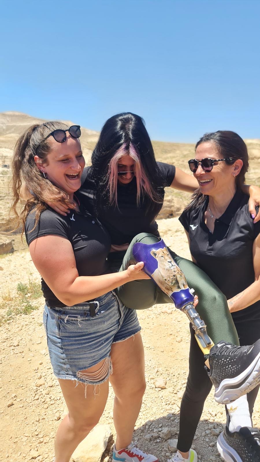Three women laughing as they carry a friend with a prosthetic leg on a desert trip
