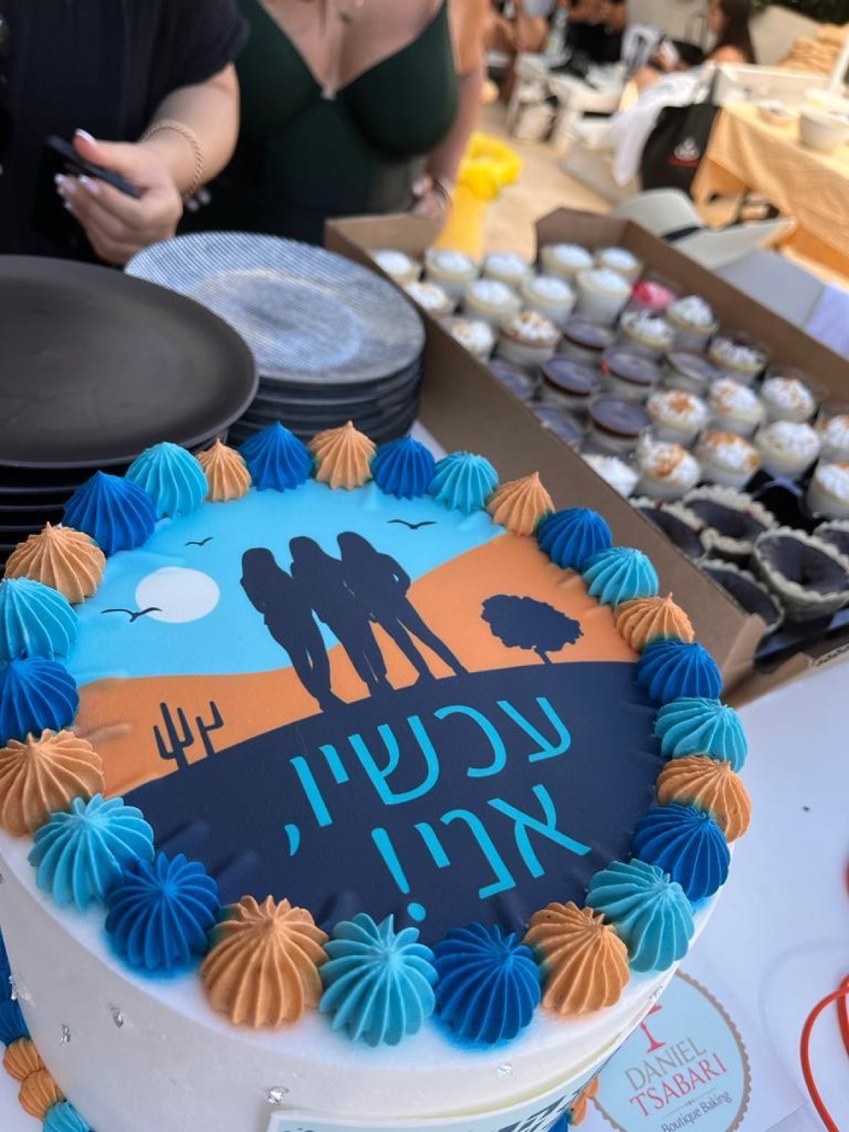 A decorated cake with the Now, Me! logo, surrounded by blue and orange frosting